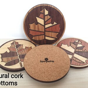 May include: Four round wooden coasters with a maple leaf design. The coasters are made of different types of wood and have a natural cork bottom. The coasters are arranged in a circle with the cork bottoms facing up. The text "SparkedUp" is visible on one of the coasters.