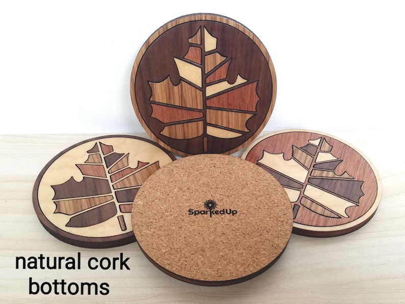 May include: Four round wooden coasters with a maple leaf design. The coasters are made of different types of wood and have a natural cork bottom. The coasters are arranged in a circle with the cork bottoms facing up. The text "SparkedUp" is visible on one of the coasters.