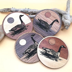 May include: Four wooden coasters featuring a black and white silhouette of a goose on each. The coasters have a natural wood finish with a dark brown stain. The geese are in different positions on each coaster.