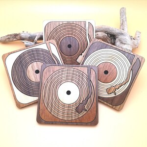 May include: Five square wooden coasters with a vinyl record player design. The coasters feature a range of wood tones, from light to dark brown, with detailed concentric circle patterns. The background includes a piece of driftwood. Each coaster is approximately 10 cm x 10 cm.