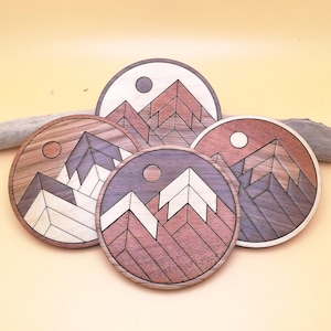 Peut inclure: Quatre sous-verres en bois avec des motifs géométriques de montagnes. Chaque sous-verre présente une palette de couleurs différente, notamment le brun, le blanc et le gris.