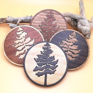 May include: Four round wooden coasters with a laser-cut design of a pine tree. The coasters are in different shades of brown and black.