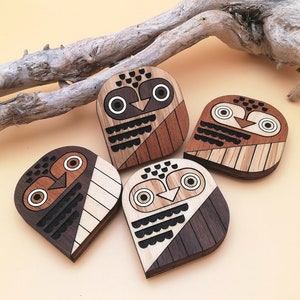 May include: Five wooden owl-shaped coasters with geometric designs. Each coaster features a different combination of light and dark wood tones, with details in black and white. The coasters are approximately 10 cm wide and 10 cm tall.