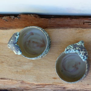 May include: Two handmade ceramic bowls with a unique design. The bowls are light brown with a speckled, textured rim and a handle-like extension. The interior has a swirl pattern. The bowls are placed on a wooden surface.