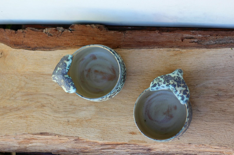 May include: Two handmade ceramic bowls with a unique design. The bowls are light brown with a speckled, textured rim and a handle-like extension. The interior has a swirl pattern. The bowls are placed on a wooden surface.