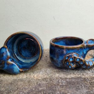 Könnte beinhalten: Zwei Keramikbecher mit tiefblauer Glasur und braunen Randakzenten. Ein Becher liegt auf der Seite, während der andere aufrecht steht und einen Griff in Form eines texturierten Oktopus-Tentakels mit sichtbaren Saugnapfdetails aufweist.