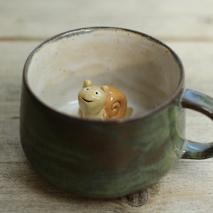 Könnte beinhalten: Eine handgefertigte Keramiktasse mit grüner und brauner Glasur. Im Inneren der Tasse befindet sich eine kleine, lächelnde Schneckenfigur. Die Tasse hat eine abgerundete Form und einen Henkel. Die Tasse steht auf einer Holzoberfläche.