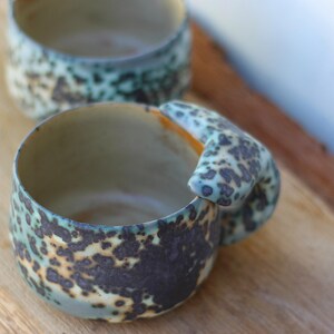 May include: Two handmade ceramic mugs with a speckled teal and brown glaze. One mug is in focus, showcasing a unique handle design. The mugs are set on a wooden surface, highlighting their rustic aesthetic. The mugs are ideal for coffee or tea.