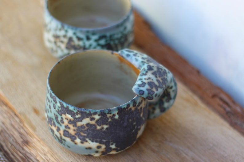 May include: Two handmade ceramic mugs with a speckled teal and brown glaze. One mug is in focus, showcasing a unique handle design. The mugs are set on a wooden surface, highlighting their rustic aesthetic. The mugs are ideal for coffee or tea.