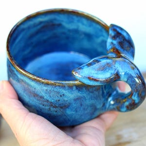 May include: A blue ceramic bowl with a whale tail handle. The bowl has a glossy finish and a brown rim. The whale tail handle is a unique design element. This bowl is ideal for serving soups, cereal, or snacks. The bowl's diameter is approximately 10 cm.