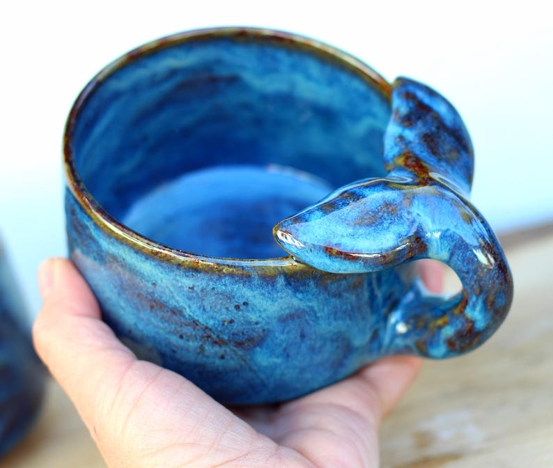 May include: A blue ceramic bowl with a whale tail handle. The bowl has a glossy finish and a brown rim. The whale tail handle is a unique design element. This bowl is ideal for serving soups, cereal, or snacks. The bowl's diameter is approximately 10 cm.
