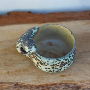 May include: A handcrafted ceramic mug with a distinctive handle. The mug has a speckled pattern in black and light green on a cream base. The handle has a unique shape and colour. The mug is placed on a wooden surface.