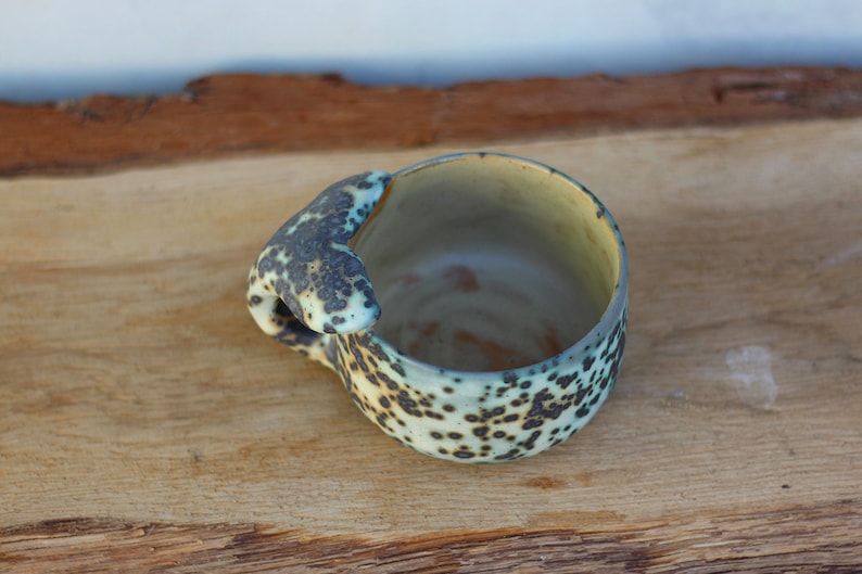 May include: A handcrafted ceramic mug with a distinctive handle. The mug has a speckled pattern in black and light green on a cream base. The handle has a unique shape and colour. The mug is placed on a wooden surface.