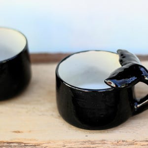 May include: Two black ceramic mugs with a whale tail handle. The mugs have a glossy finish and a white interior. The mugs are on a light-coloured wooden surface.