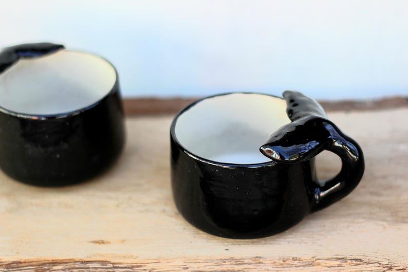 May include: Two black ceramic mugs with a whale tail handle. The mugs have a glossy finish and a white interior. The mugs are on a light-coloured wooden surface.