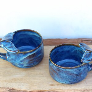 May include: Two handmade ceramic mugs with a whale tail handle design. The mugs are glazed in shades of blue with brown accents around the rim and handle. They are placed on a wooden surface.