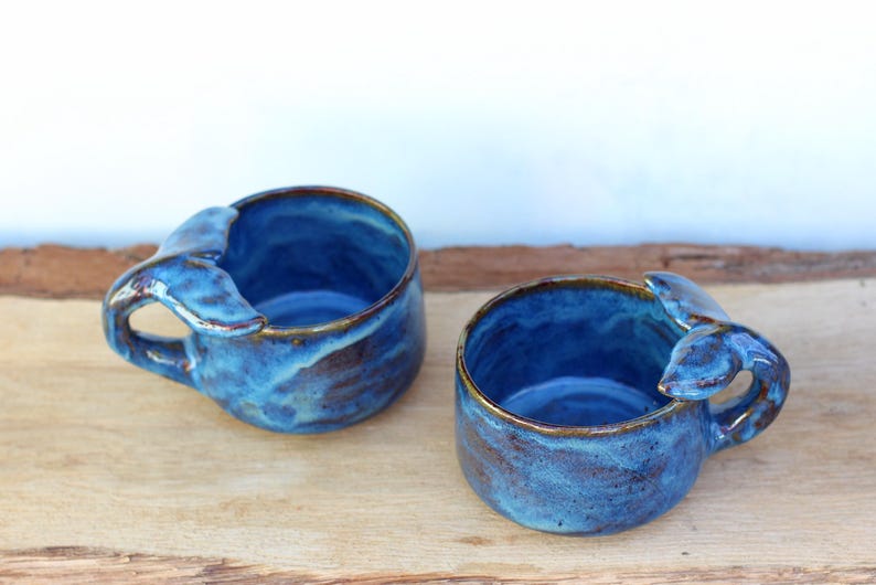 May include: Two handmade ceramic mugs with a whale tail handle design. The mugs are glazed in shades of blue with brown accents around the rim and handle. They are placed on a wooden surface.