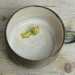 Könnte beinhalten: Eine Keramiktasse mit grün-brauner Glasur, mit einer kleinen, gelb-grünen Raupenfigur im Inneren. Die Tasse hat eine runde Form und einen Henkel, ideal für ein warmes Getränk.