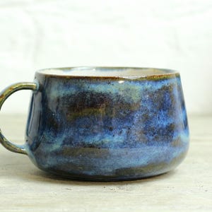 Könnte beinhalten: Eine handgefertigte Keramik-Tasse mit abgerundeter Form und einer dunkelblauen Glasur mit helleren blauen und braunen Akzenten. Die Tasse hat einen kleinen, abgerundeten Henkel und einen hellfarbigen Rand. Die Tasse steht auf einer hellen Holzoberfläche.