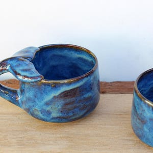 May include: Two handmade ceramic mugs with a whale tail handle design. The mugs are glazed in shades of blue with brown accents, giving them a unique, artistic appearance. The mugs are sitting on a wooden surface.