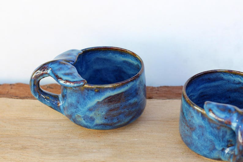 May include: Two handmade ceramic mugs with a whale tail handle design. The mugs are glazed in shades of blue with brown accents, giving them a unique, artistic appearance. The mugs are sitting on a wooden surface.