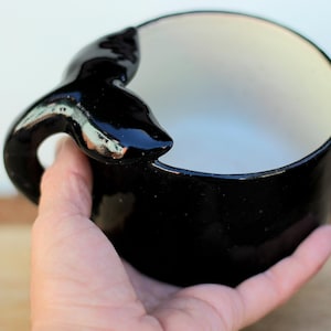 May include: A black ceramic bowl with a whale tail handle. The interior is white, contrasting with the glossy black exterior. The whale tail handle adds a unique, decorative element to the bowl's design.