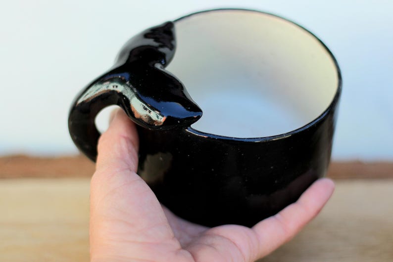 May include: A black ceramic bowl with a whale tail handle. The interior is white, contrasting with the glossy black exterior. The whale tail handle adds a unique, decorative element to the bowl's design.