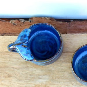 May include: Two handmade ceramic bowls with a deep blue glaze. One bowl has a handle shaped like a bird's wing. The bowls are on a wooden surface with a natural edge. The bowls are suitable for serving dips or small portions.