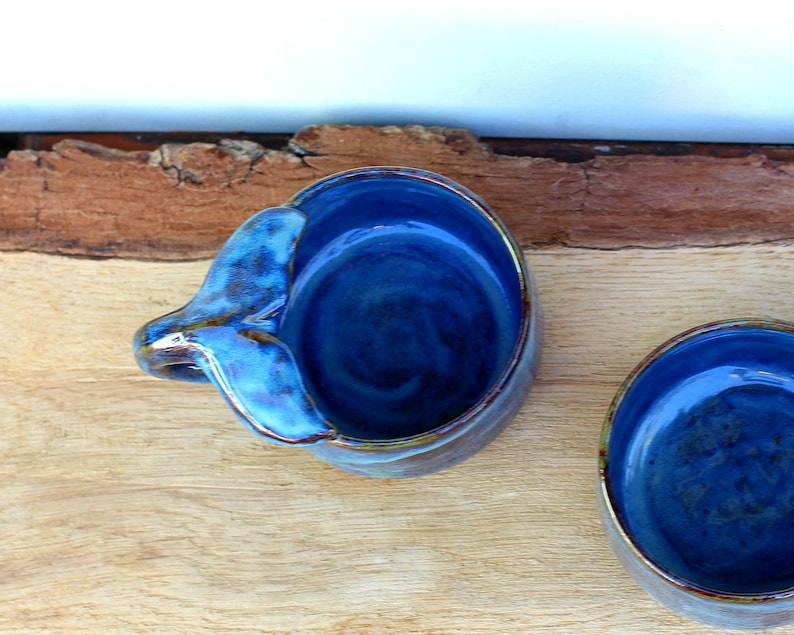 May include: Two handmade ceramic bowls with a deep blue glaze. One bowl has a handle shaped like a bird's wing. The bowls are on a wooden surface with a natural edge. The bowls are suitable for serving dips or small portions.