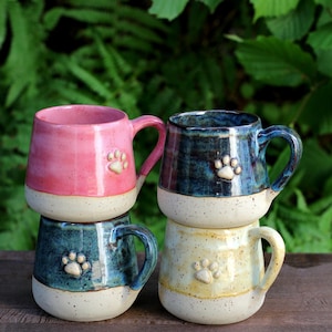 Könnte beinhalten: Vier gestapelte Keramikbecher, jeder mit einem einzigartigen Farb- und Pfotenabdruck-Design. Die Becher sind rosa, dunkelblau, blaugrün und gelb, mit einem gesprenkelten weißen Boden. Die Pfotenabdrücke sind auf der Vorderseite jedes Bechers geprägt.