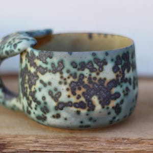 May include: A handmade ceramic mug with a distinctive handle. The mug has a speckled pattern in shades of blue, green, and brown. The interior is a light yellow colour. The mug is designed for hot drinks.