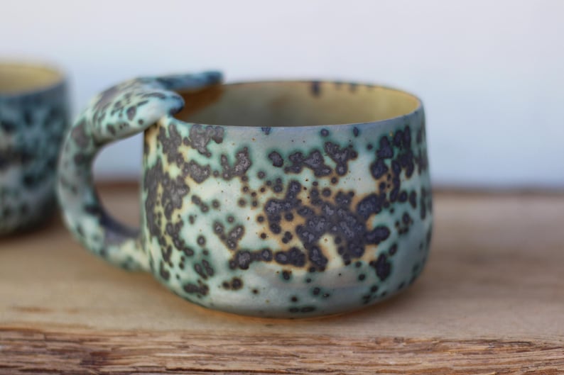 May include: A handmade ceramic mug with a distinctive handle. The mug has a speckled pattern in shades of blue, green, and brown. The interior is a light yellow colour. The mug is designed for hot drinks.