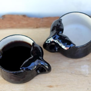 Könnte beinhalten: Zwei schwarze Keramikbecher mit Walflossendesign. Ein Becher ist mit einer dunklen Flüssigkeit gefüllt, der andere ist leer. Die Becher haben eine weiße Innenseite und stehen auf einer Holzoberfläche.