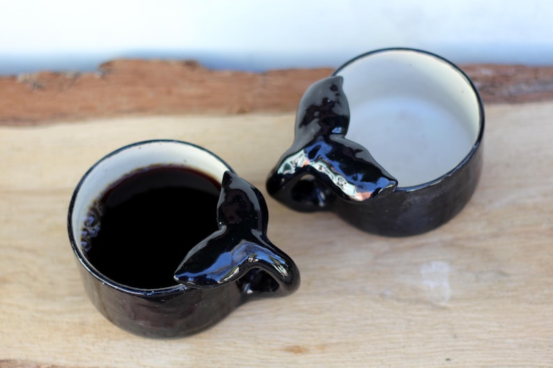 May include: Two black ceramic mugs with a whale tail design. One mug is filled with a dark liquid, the other is empty. The mugs have a white interior and sit on a wooden surface.