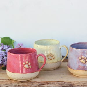 Könnte beinhalten: Drei handgefertigte Keramikbecher in Rosa, Gelb und Lavendel, jeweils mit einem Pfotenabdruck-Design. Die Becher haben ein zweifarbiges Farbschema mit hellbeigem Sockel. Ein Zweig lila Blumen befindet sich im Hintergrund.