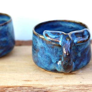 May include: Two ceramic mugs with a whale tail handle. The mugs are a vibrant blue with brown accents and a glossy finish. The mugs are on a wooden surface.