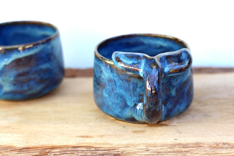 May include: Two ceramic mugs with a whale tail handle. The mugs are a vibrant blue with brown accents and a glossy finish. The mugs are on a wooden surface.