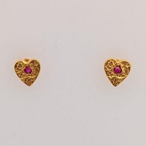 Puede incluir: Un par de pendientes de botón dorados en forma de corazón. Cada pendiente presenta una superficie texturizada y una pequeña gema roja redonda en el centro, sobre un fondo blanco.