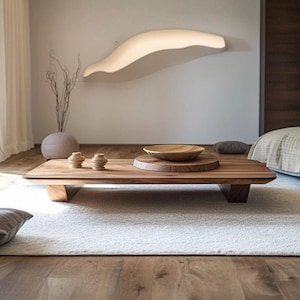 May include: A low-profile rectangular wooden coffee table with a natural grain finish, featuring two small wooden decorative objects and a large wooden bowl placed on a circular wooden base, set upon a textured white area rug in a room.