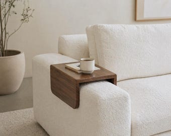 May include: A dark wood, C-shaped sofa arm table is attached to a white, textured sofa arm. A small, white and tan mug sits on the table, along with a small book. The sofa is in a neutral-toned room with a potted plant.