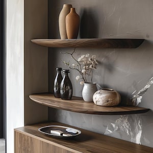 May include: Two wooden shelves with rounded edges, mounted on a grey wall. The top shelf has two ceramic vases, one brown and one beige. The bottom shelf has two glass bottles, a white ceramic vase, and a brown ceramic bowl. Dried flowers are in the white vase.