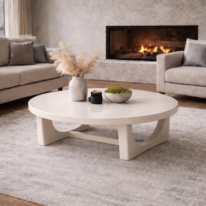 May include: A round, off-white coffee table with a glossy finish. The table has a unique base design and sits on a patterned rug. Decorative items, including a vase with dried plants, are displayed on the table.