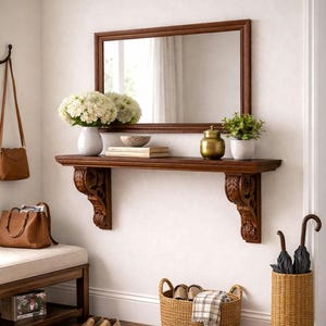 Könnte beinhalten: Ein dunkel gerahmter Spiegel hängt über einem passenden Regal mit geschnitzten Konsolen. Auf dem Regal stehen eine weiße Vase mit weißen Blumen, eine kleine Schale, Bücher, ein goldener Topf und eine Topfpflanze. Eine Bank, ein Korb und ein Regenschirmständer vervollständigen den Eingangsbereich.