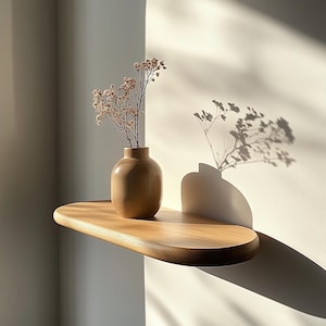 Puede incluir: Una estantería de madera con bordes redondeados, montada en una pared blanca. Un pequeño jarrón de madera con flores secas se encuentra sobre la estantería. La estantería y el jarrón están iluminados por la luz del sol.