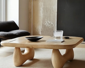 May include: A light-colored wooden coffee table with a unique, organic shape. The table has a smooth, flat surface and is supported by four curved, sculptural legs. A black bowl, a glass, and a book are on the table.