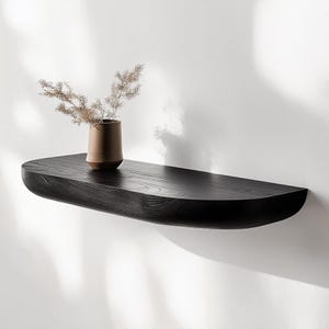 May include: A black wooden shelf with a curved edge, mounted on a white wall. A small brown ceramic vase with dried flowers sits on the shelf.