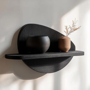 Könnte beinhalten: Modernes, schwarzes Wandregal aus Holz mit einer einzigartigen, organischen Form. Es hat ein abgerundetes Regal und eine dekorative Rückwand. Zwei dekorative Vasen stehen auf dem Regal: eine dunkle, runde Vase und eine kleinere, beige Vase mit getrocknetem Laub.