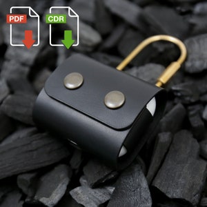 May include: A black leather case with a gold metal clasp and two silver buttons. The case is designed to hold a pair of wireless earbuds. The case is sitting on a bed of black charcoal.