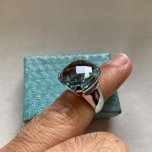 May include: A large, faceted, light blue gemstone ring with a silver band. The ring is set on a finger, with a small, patterned, blue box in the background. The gemstone has a square shape.