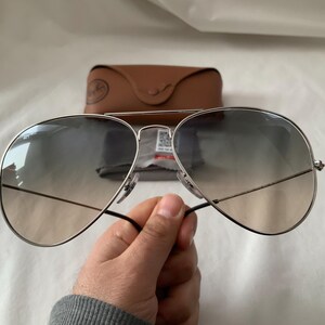 May include: Silver-framed aviator sunglasses with gradient lenses. The sunglasses have a double bridge and black temples. A brown leather case and cleaning cloth are in the background.